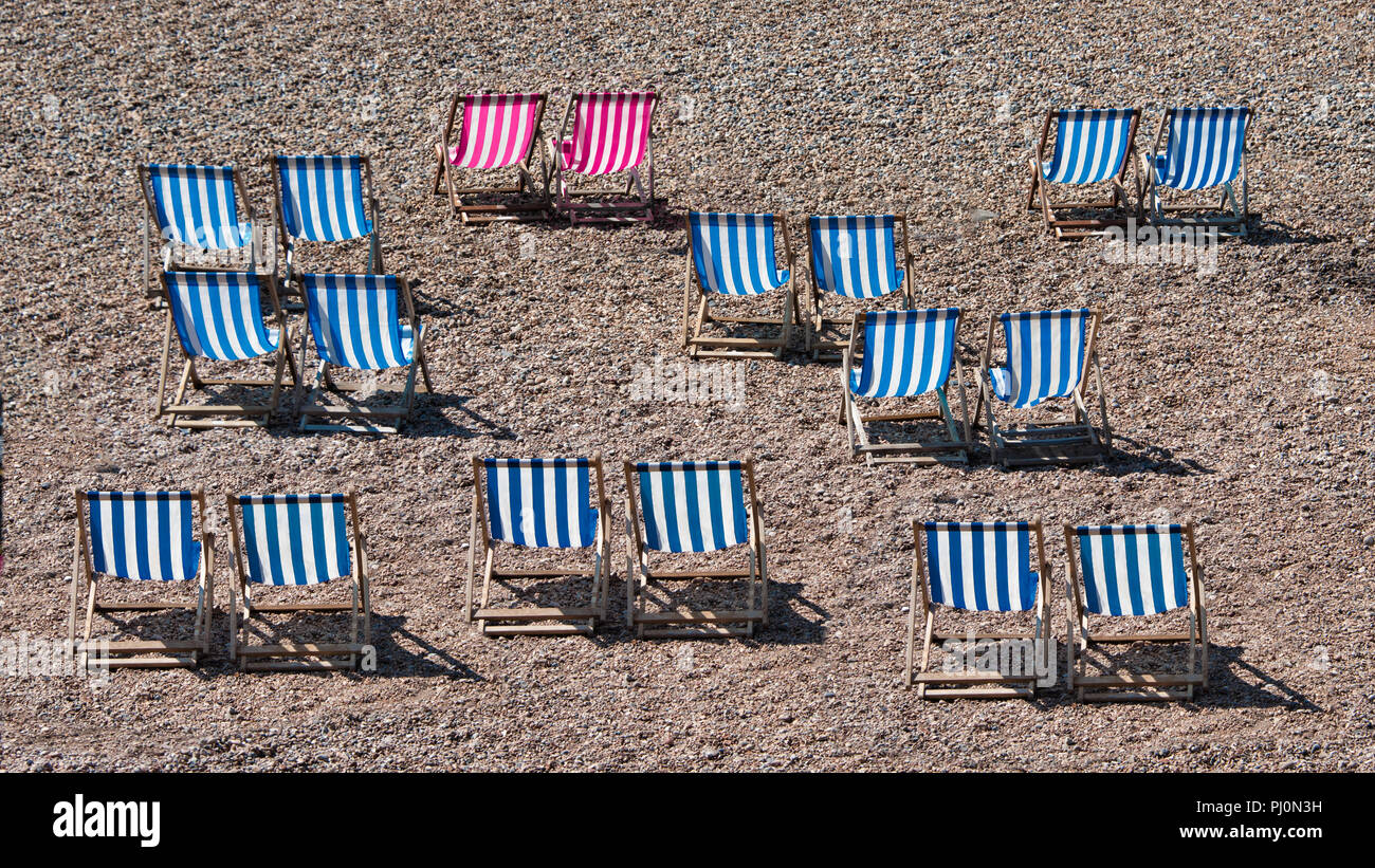 Deckchairs lined up on a pebble beach at the seaside. There are no people in the photograph. Two of the striped chairs are a different colour Stock Photo