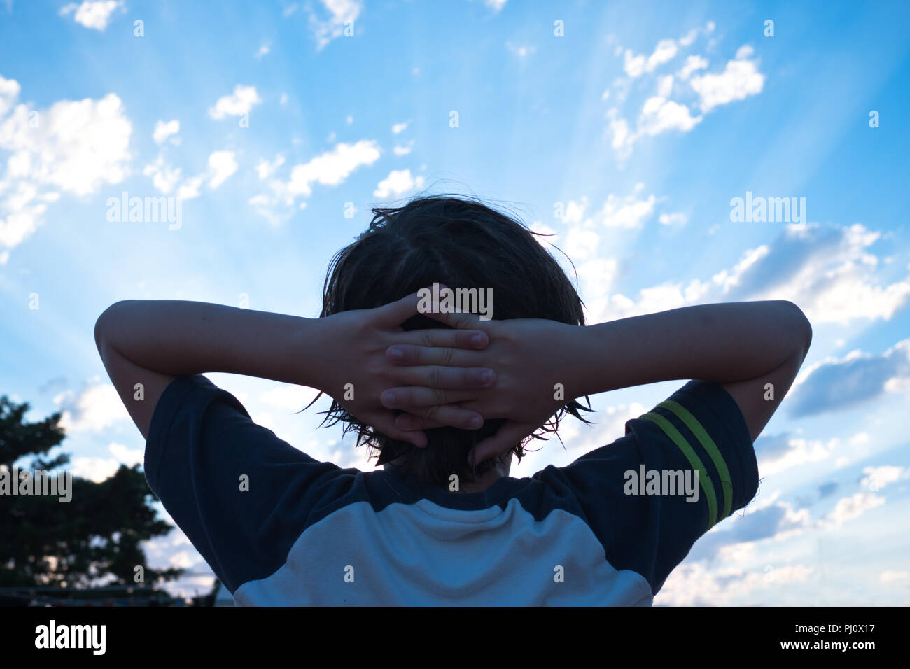 rear view of 11 year old boy with his arms outstretched and his head resting in his hands, looking up at the sky during sunset in Menorca, Spain Stock Photo