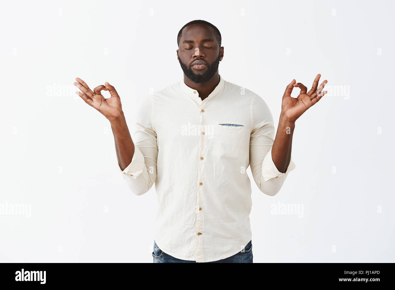 Reuniting with anture through meditation. Calm good-looking african american male in white casual shirt, raising hands in zen gesture, closing eyes and standing with calm expression over grey wall Stock Photo