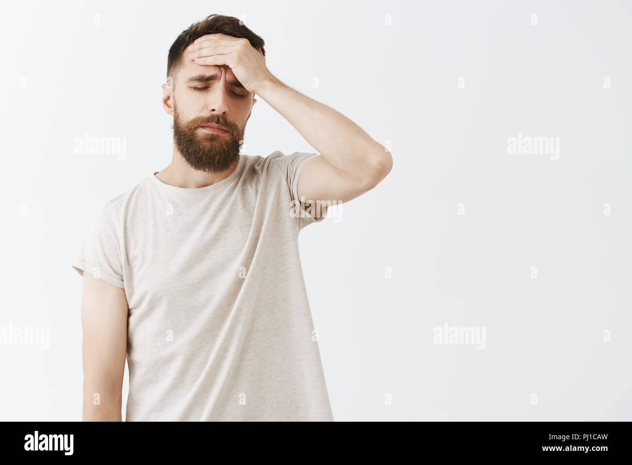 Studio shot of man feeling regret after losing during competition feeling upset and concerned holding hand on forehead closing eyes and sighing standing exhausted and tired over gray wall Stock Photo