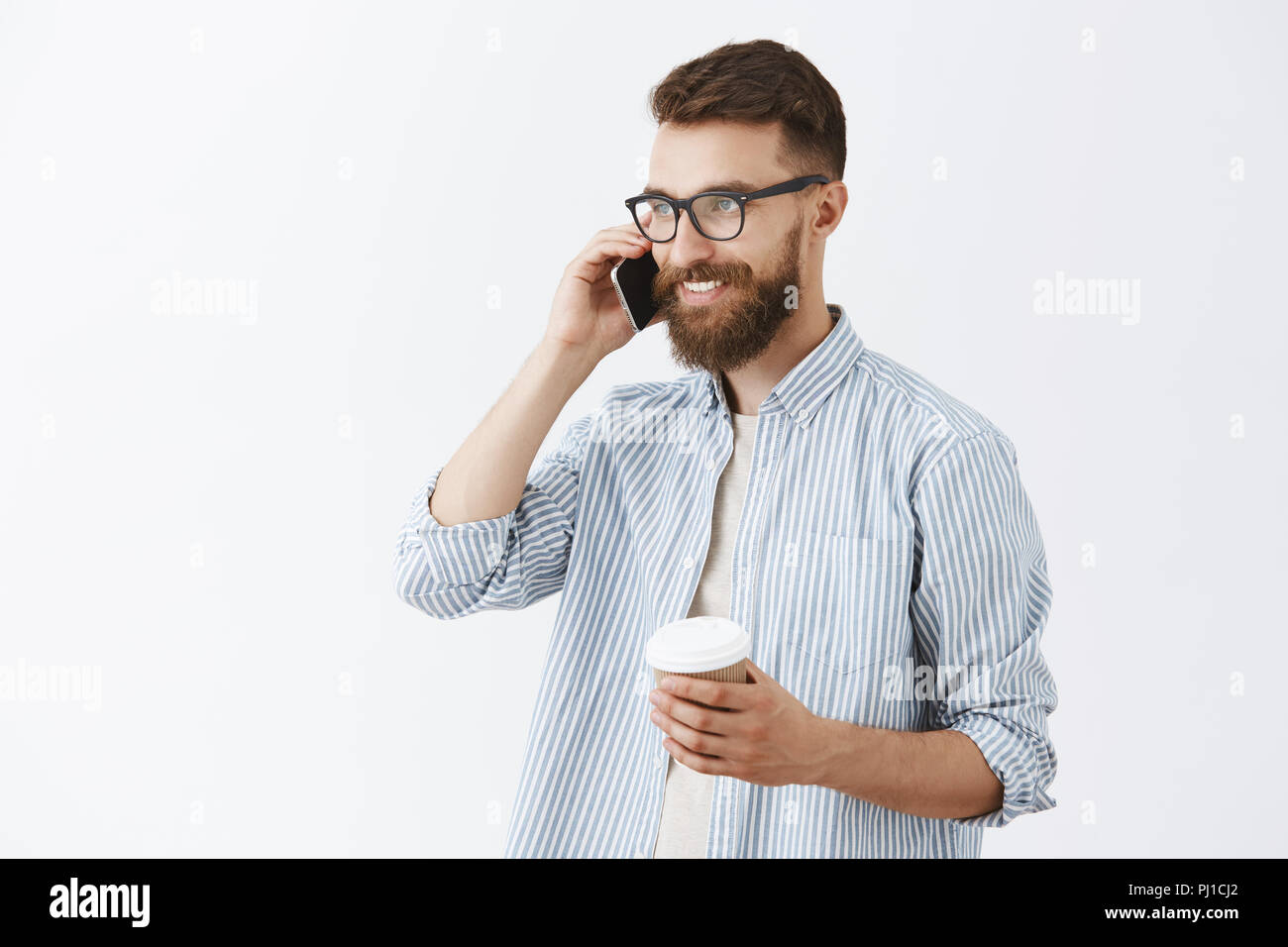 Stylish creative businessman with long beard and cool moustache talking on smartphone turning left smiling with delighted smile having great deal with business partner drinking coffee from paper cup Stock Photo