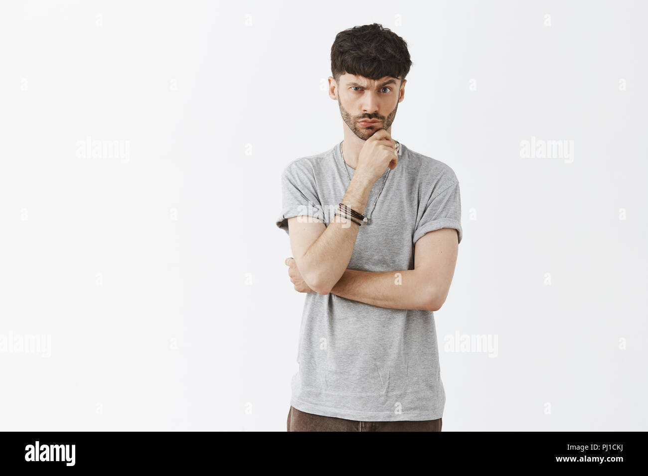 Man having disbelief listening suspicious guy giving speech. Portrait of intense doubtful good-looking urban guy with dark hair lifting eyebrow touching chin and pursing lips over gray wall Stock Photo