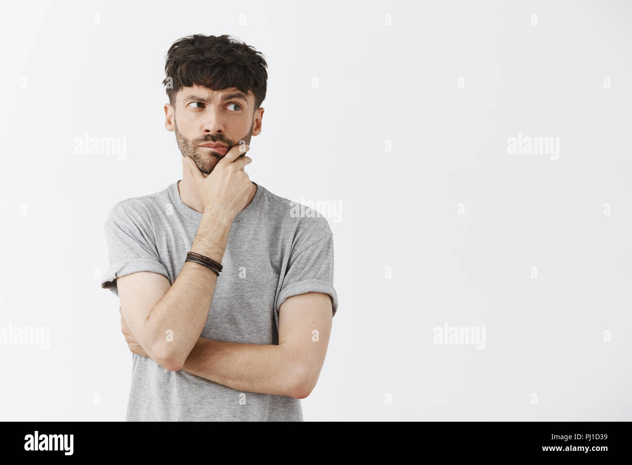 Waist-up shot of creative and artistic caucasian male model with beard holding hand on jaw and chin gazing right with spaced out expression thinking making plan over gray background Stock Photo