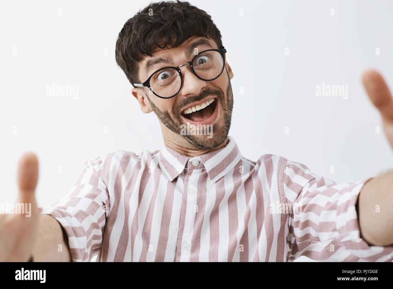 Close-up shot of funny emotive and happy good-looking modern male hipster in glasses and pink striped shirt pulling hands towards camera to make selfie, aping and making faces, fooling around Stock Photo