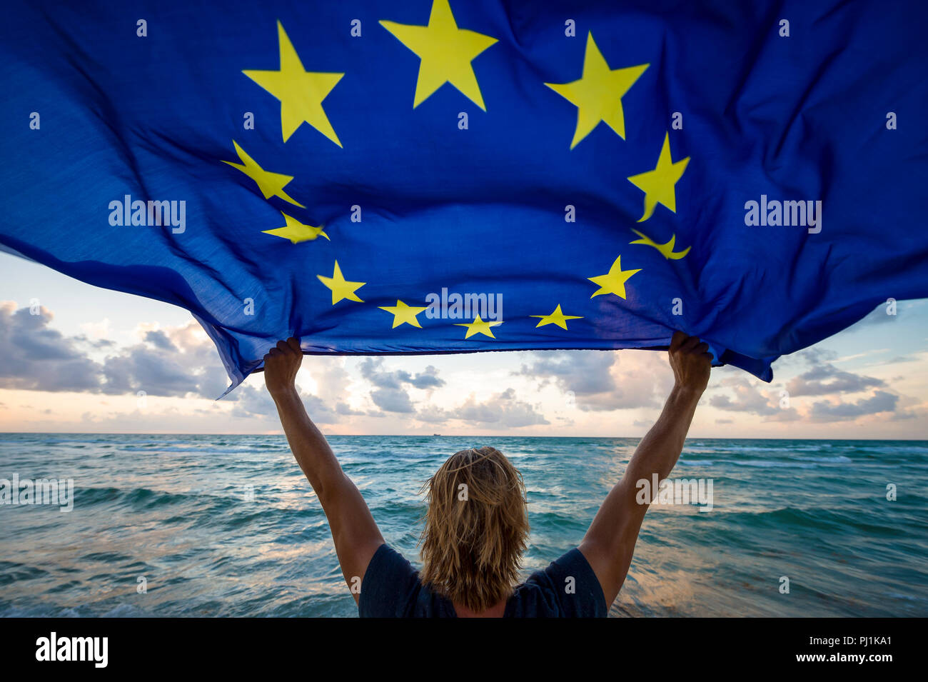 Man holding a fluttering iconic EU flag with circle of stars on beach at sunrise Stock Photo