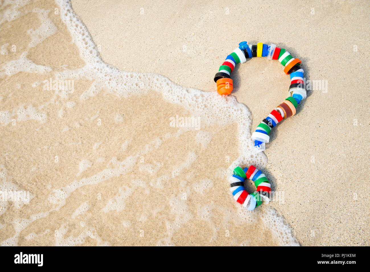 Colorful bottle caps in the shape of a question mark on a sandy beach as a wave washes up. Take action on pollution - reduce, reuse and recycle Stock Photo