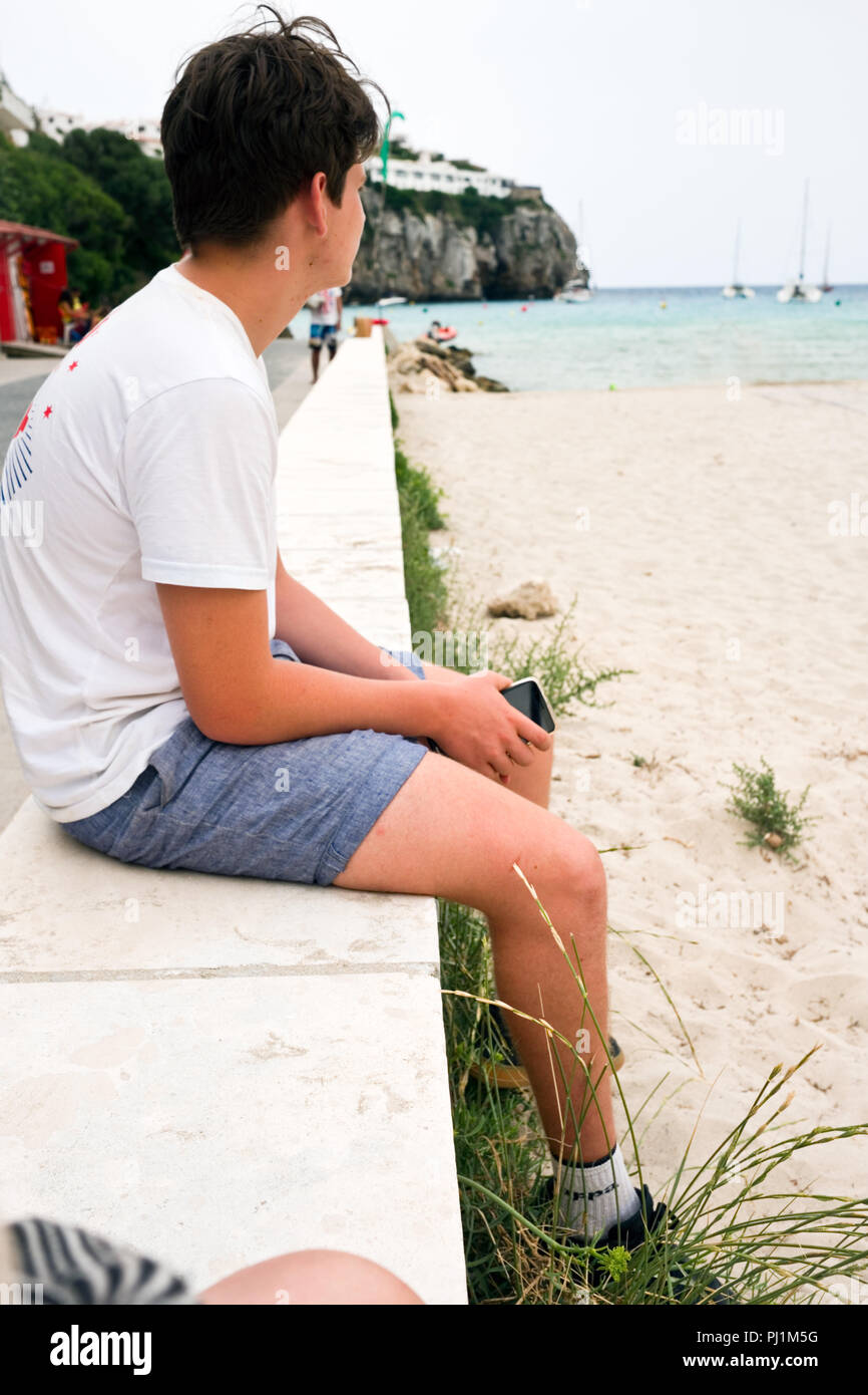 cropped view of male caucasian teenager with white tshirt and denim shorts sitting on a wall by the beach and sea holding his smartphone in his hands Stock Photo