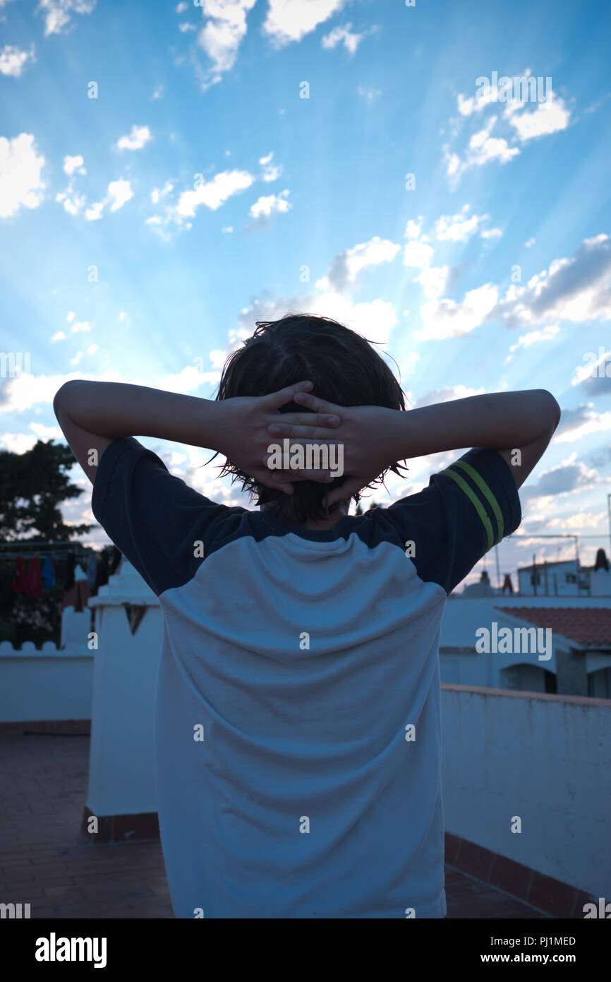 rear view of 11 year old boy with his arms outstretched and his head resting in his hands, standing on a roof terrace in Menorca, looking at a sunset Stock Photo