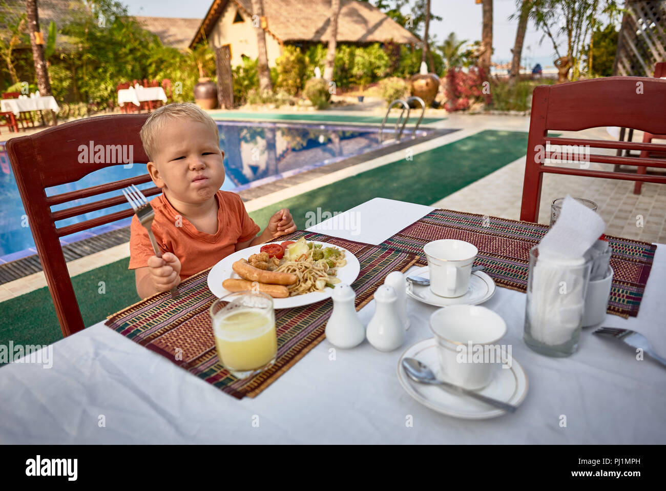 Cute boy picky eater frustrating over the dish with sausages vegetables spaghetti served with fresh pineapple juice on the table by the swimming pool Stock Photo