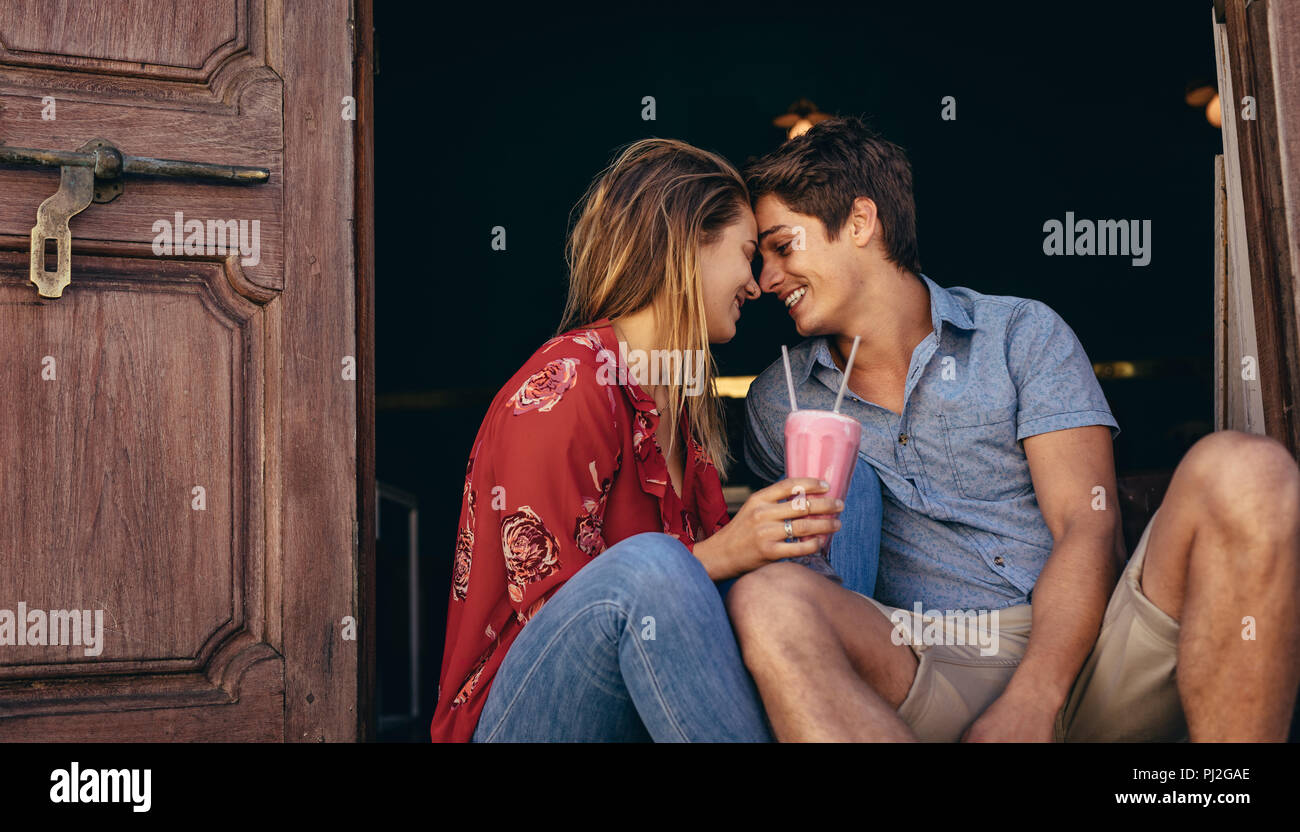 Romantic couple sitting together touching their heads. Smiling man and woman sitting at the entrance of a restaurant holding milkshake with two straws Stock Photo