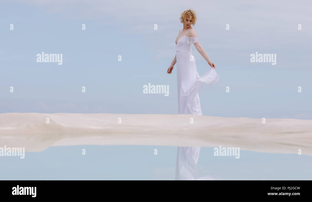 Woman in white gown standing at the beach with her reflection in water. Stock Photo