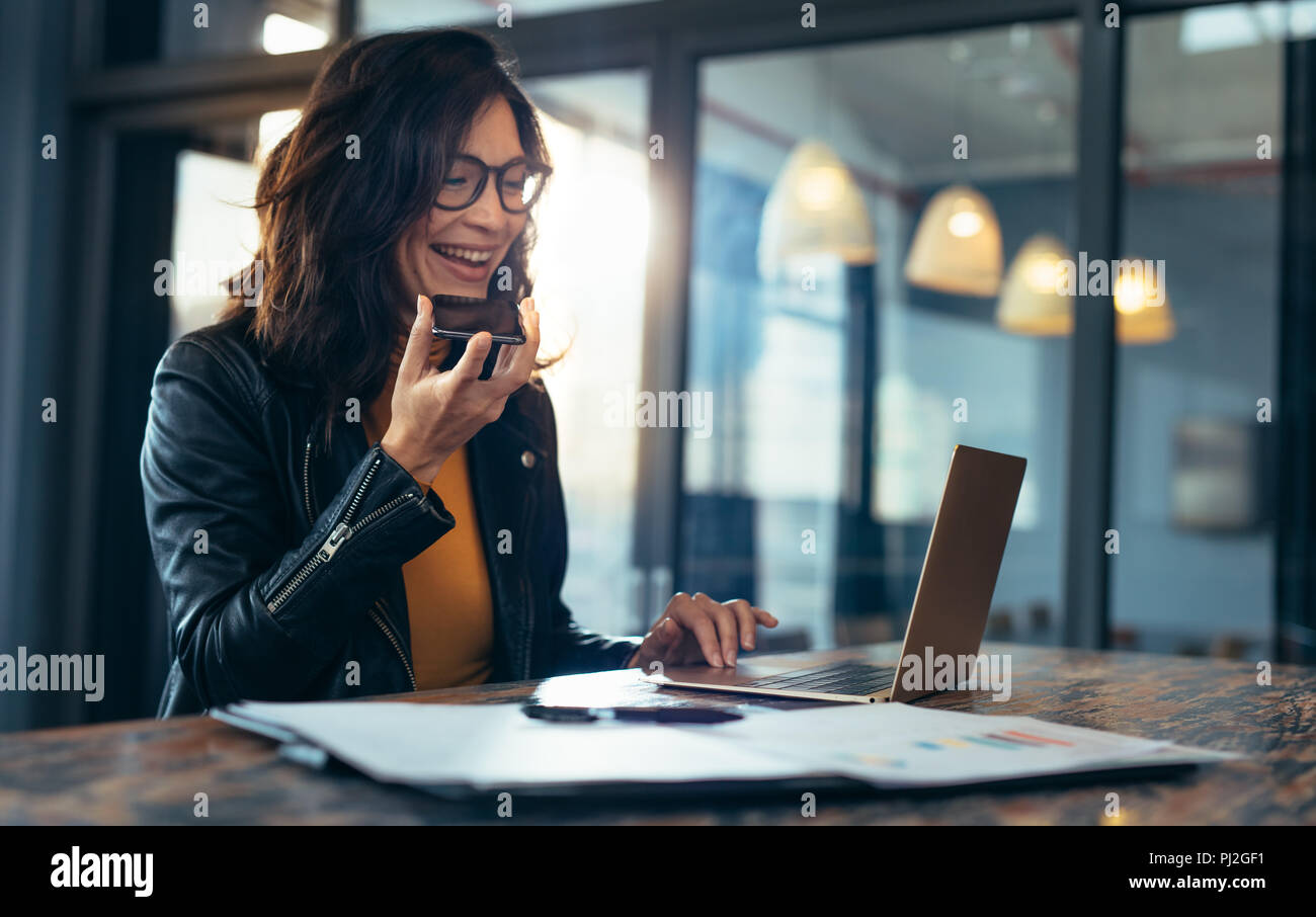 Happy business woman talking on cell phone sitting at table and working on laptop. Asian female sitting at office having telephonic conversation with  Stock Photo