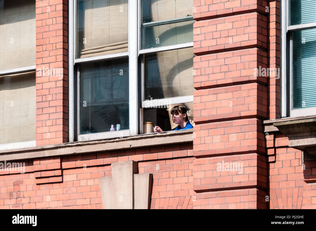 Female worker taking a cigarette break and looking out of a window from a building in Manchester Stock Photo