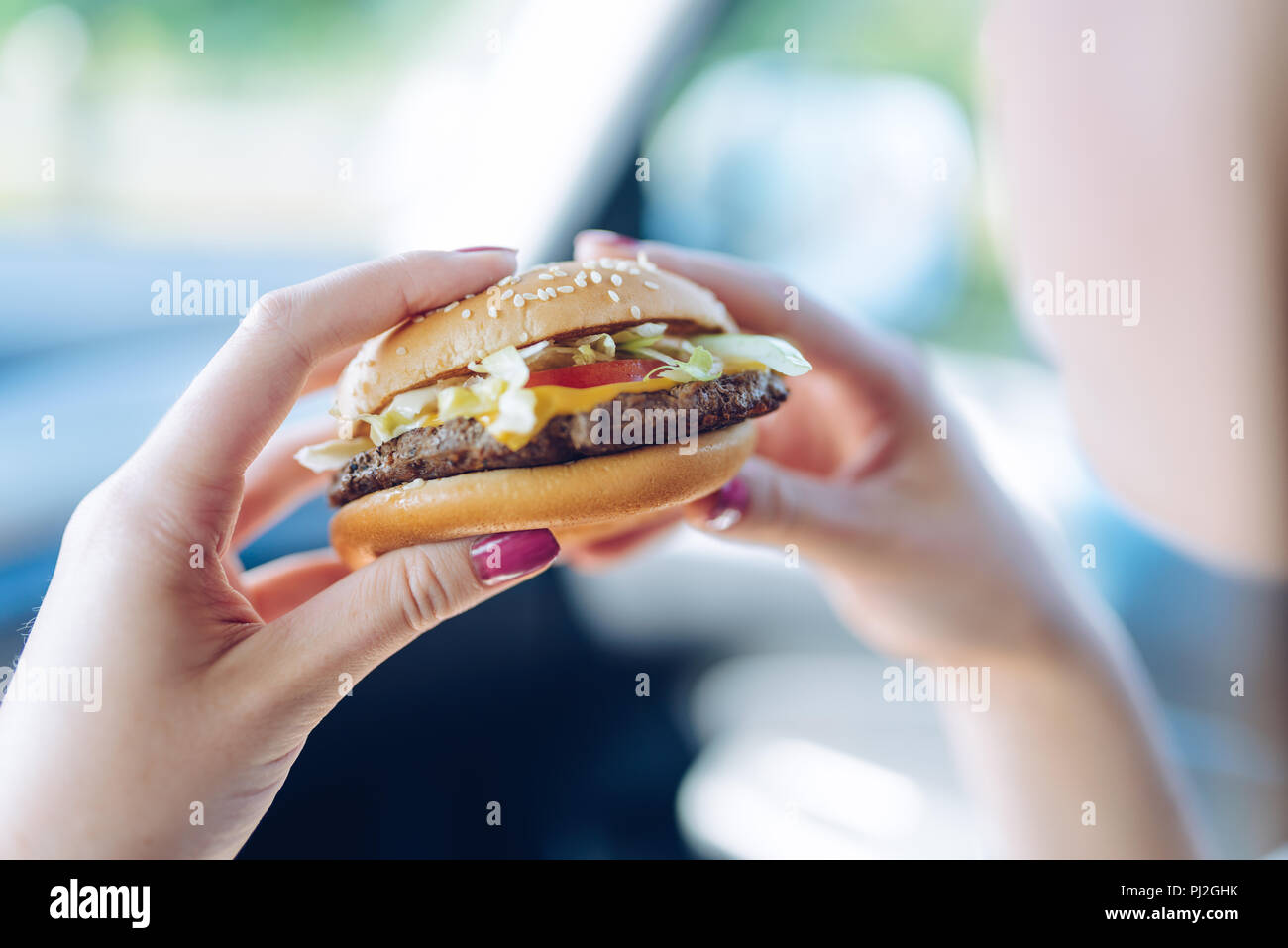 Girl holding a hamburger in  her hands sitting in a car. Unhealthy eating concept - shallow depth of field Stock Photo