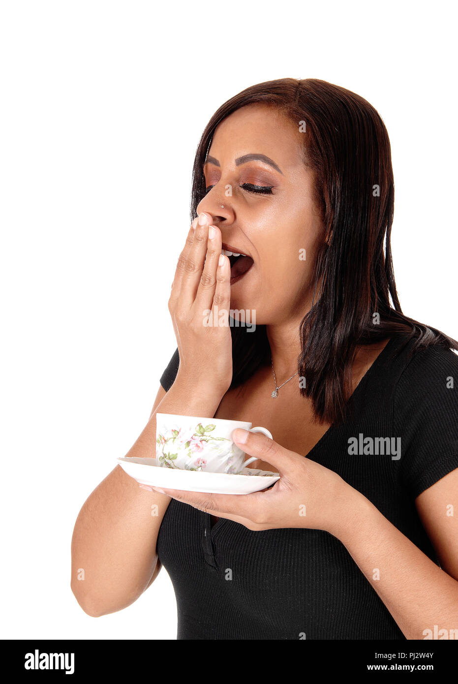 A beautiful young woman holding a coffee cup in her hand and yawning with her hand over her mouth, isolated for white background Stock Photo