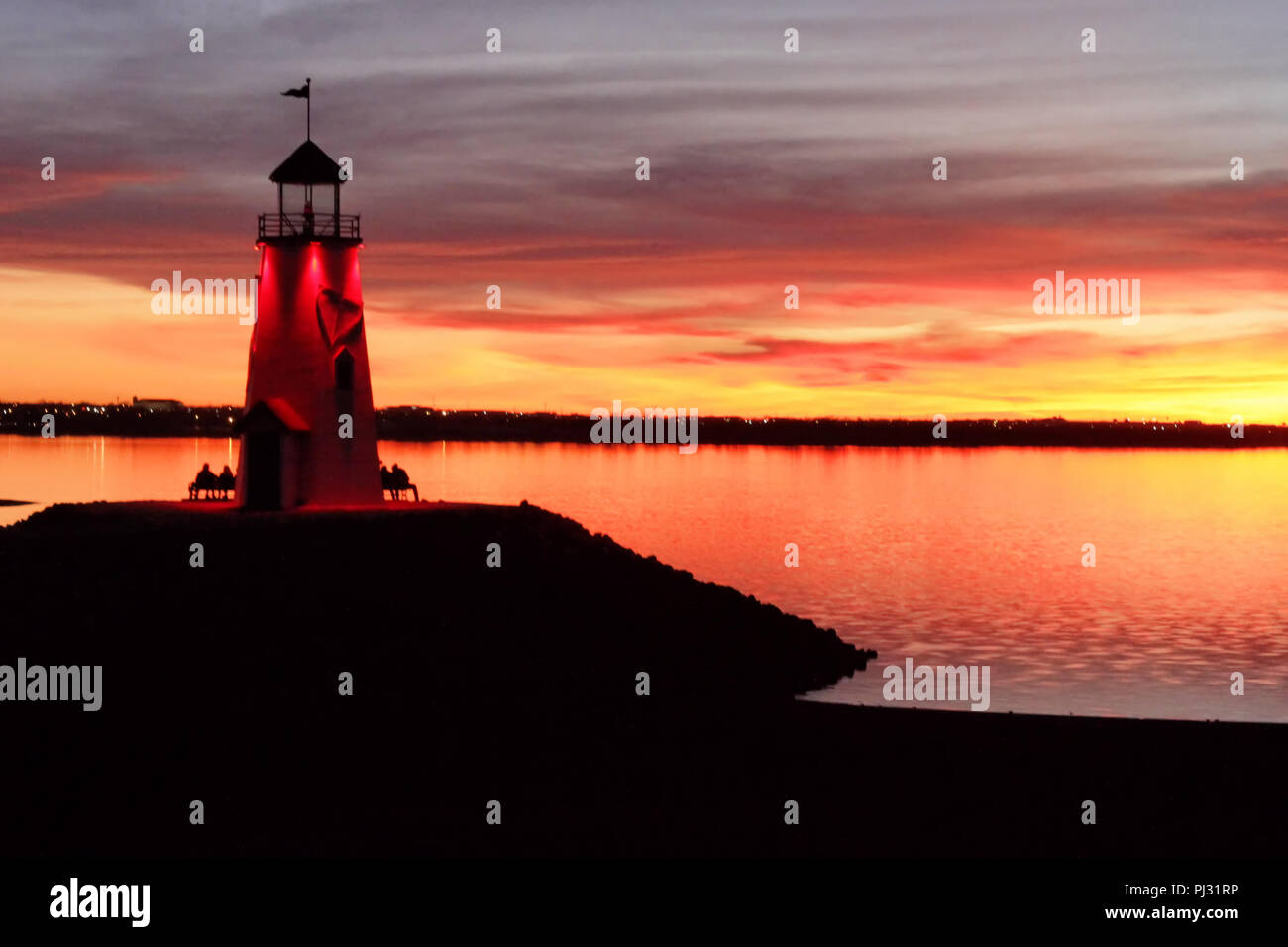 The lighthouse at Lake Hefner's East Wharf in Oklahoma City is lit up red against a colorful cloudy sunset, with people on benches. Stock Photo