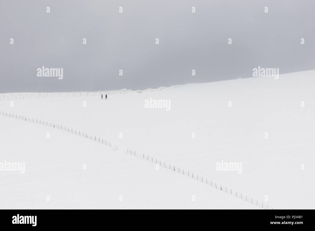 A very minimalistic view of two distant people over a mountain covered by snow, near a fence Stock Photo