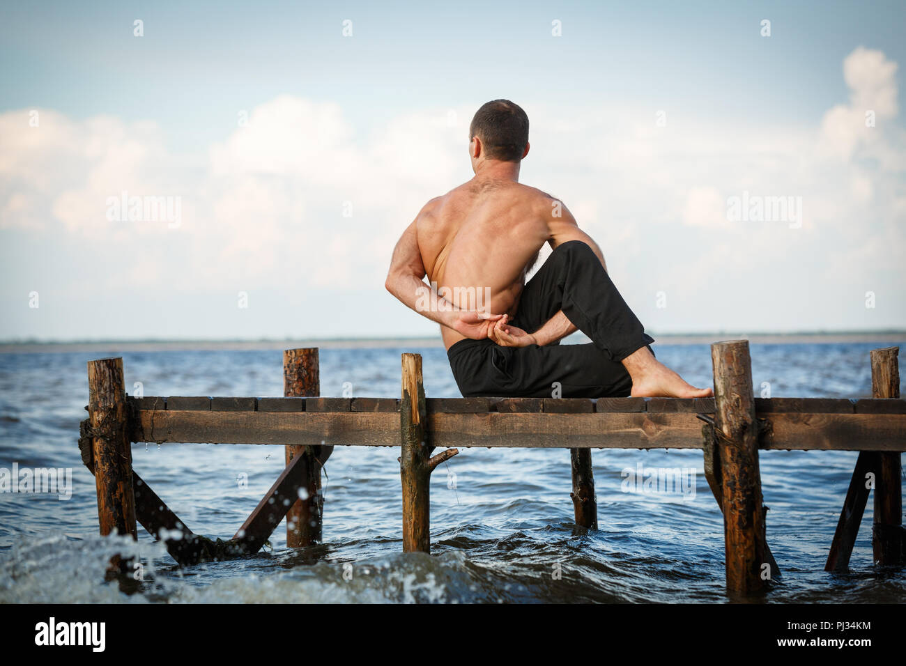 Young yoga trainer practicing ardha matsyendrasana or lord of the fishes pose on a wooden pier on a sea or river shore. Healthy lifestyle concept Stock Photo
