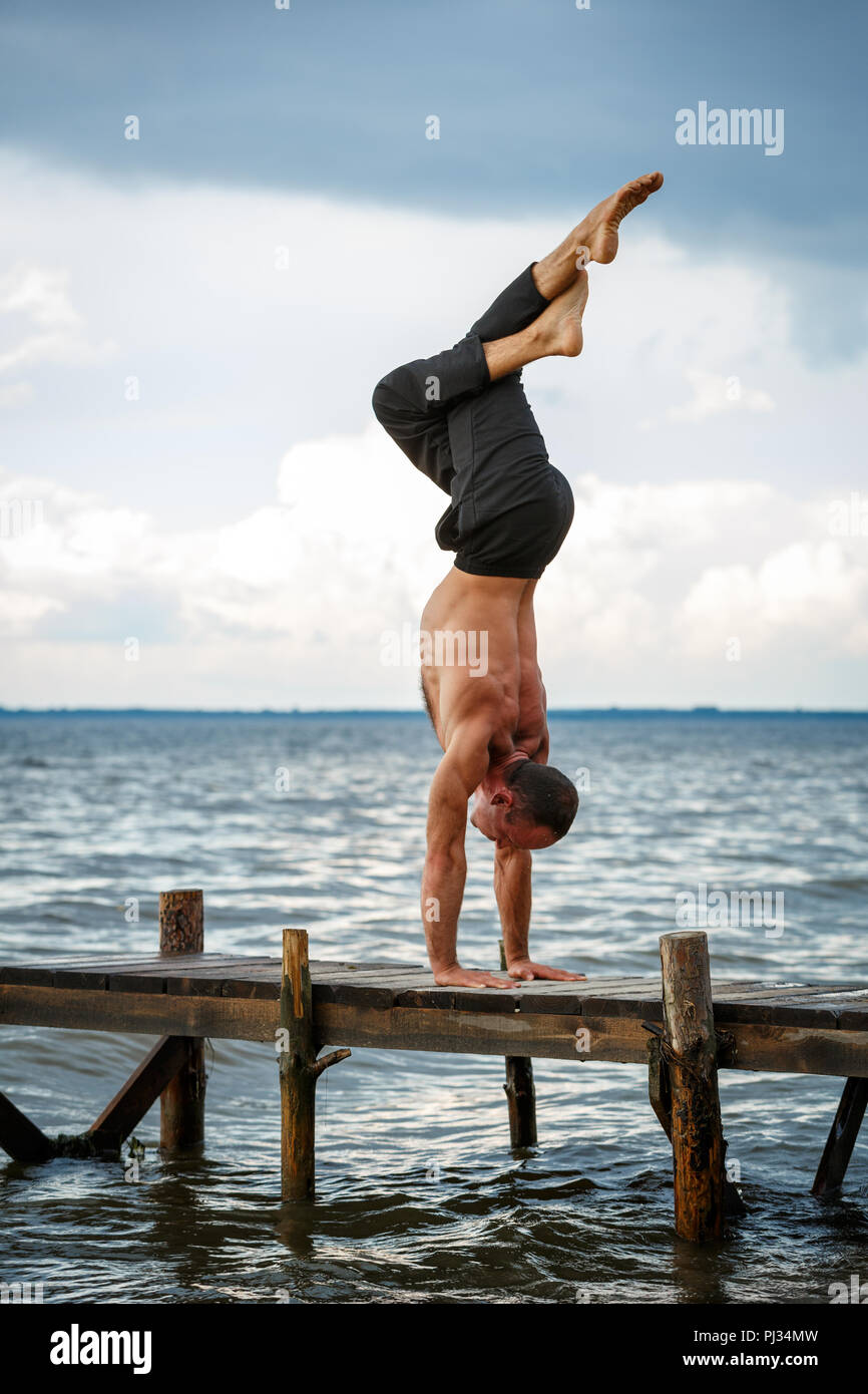 Young yoga trainer practicing handstand with legs crossed on a wooden pier on a sea or river shore. Healthy lifestyle concept Stock Photo