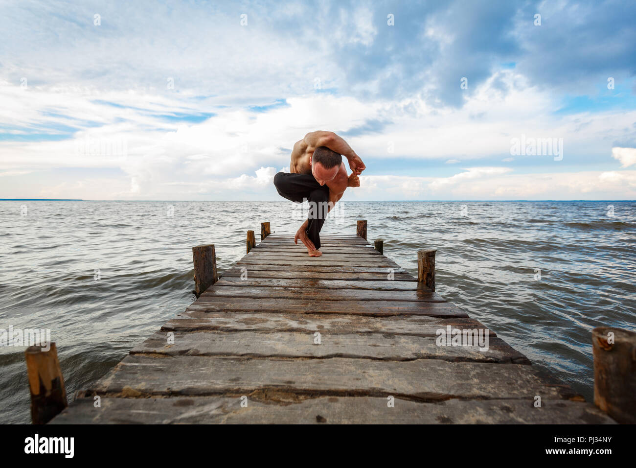 Young yoga trainer practicing yoga exercises on a wooden pier on a sea or river shore with a scenic view. Healthy lifestyle concept Stock Photo