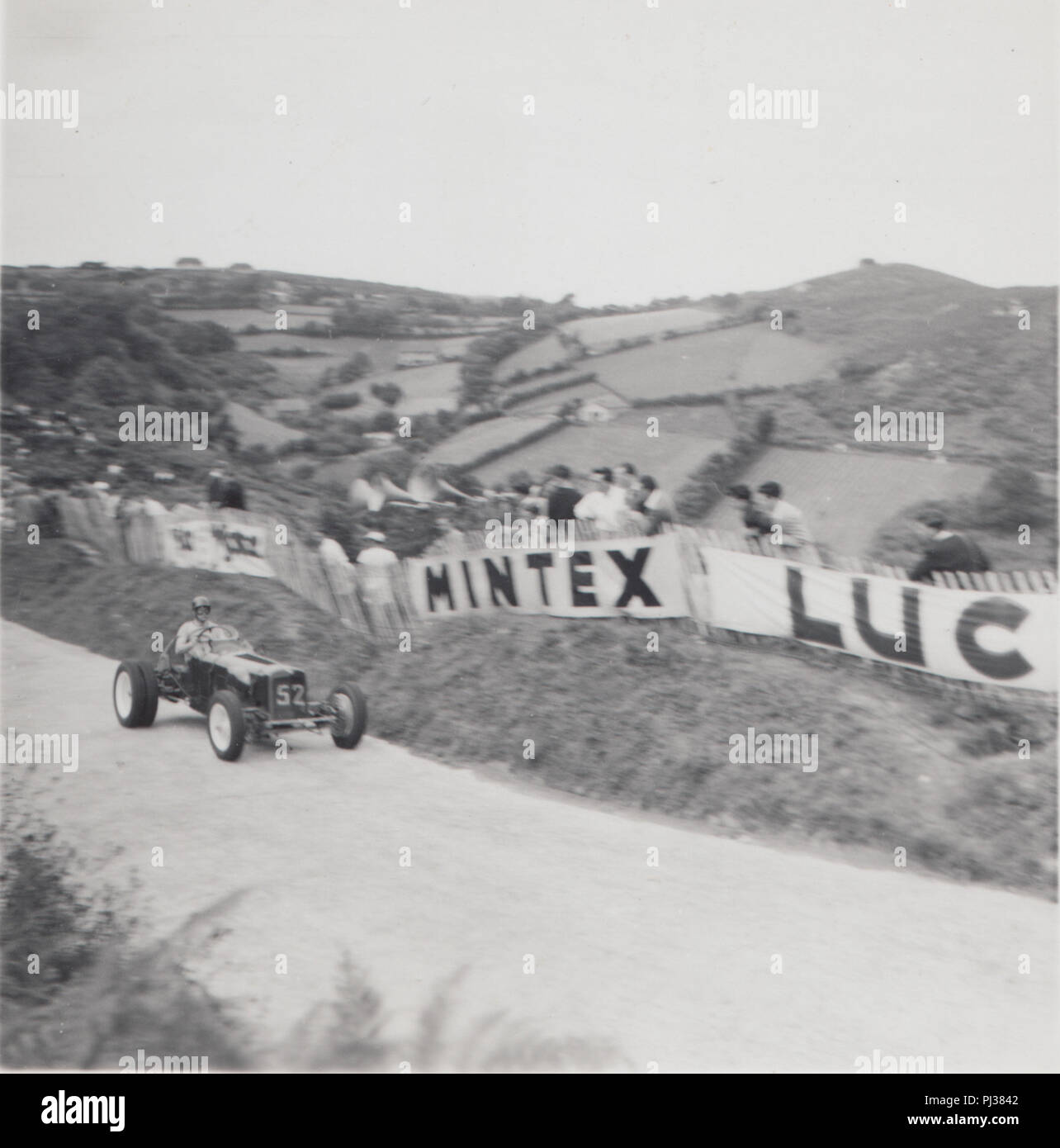 Vintage Photograph of a British Motor Racing Car With Spectators Lining The Roadside. Mintex Luc Barrier Signage. Stock Photo
