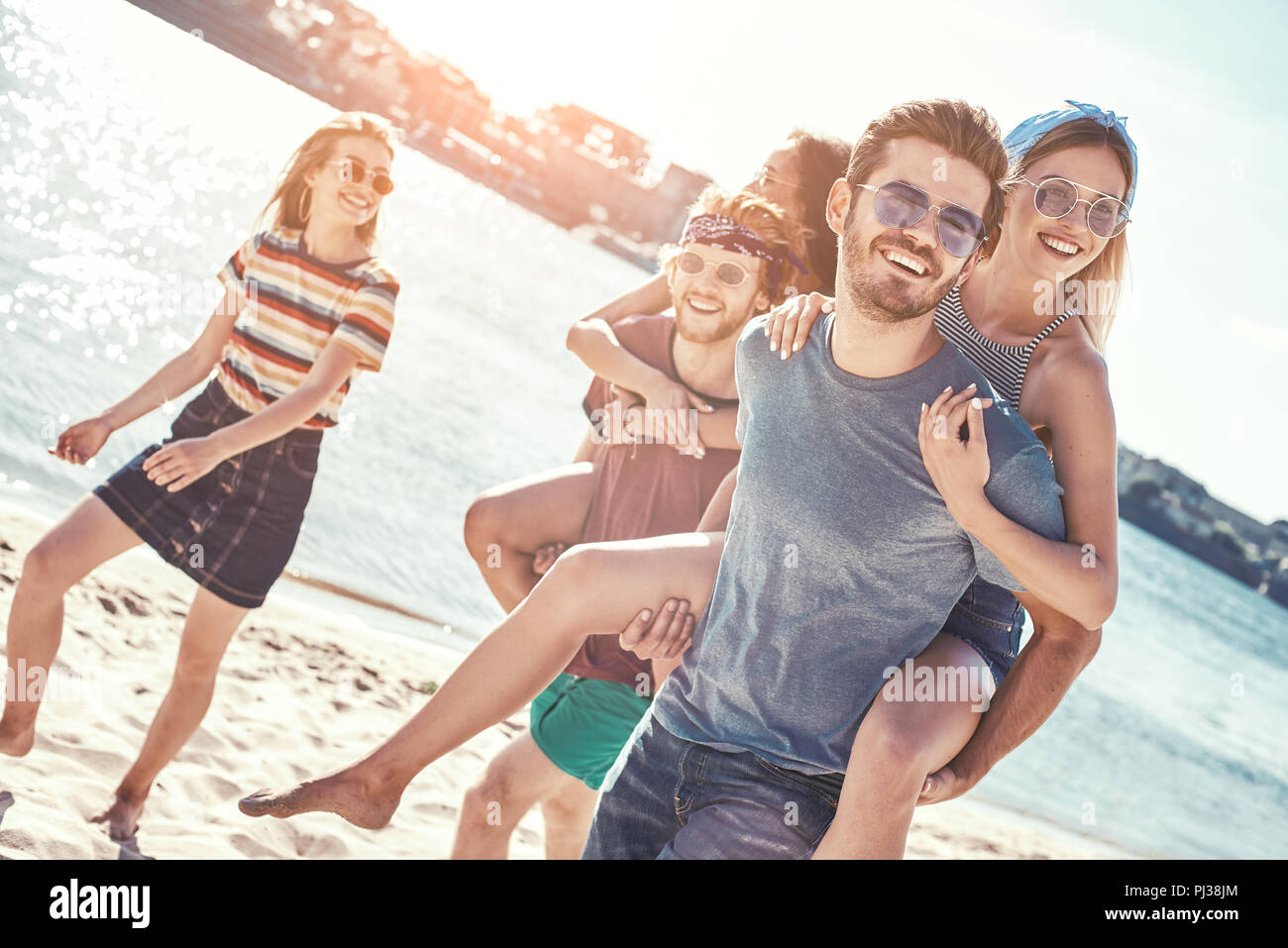 Outdoors photo of happy boyfriends piggybacking their girlfriends at sunset on beach Stock Photo
