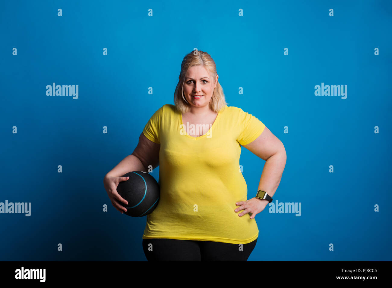 Portrait of a happy overweight woman with smartwatch and heavy ball in studio. Stock Photo