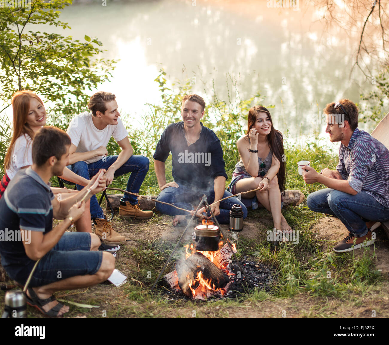 Group of friends are sitting around camp fire and preparing sausages. Stock Photo