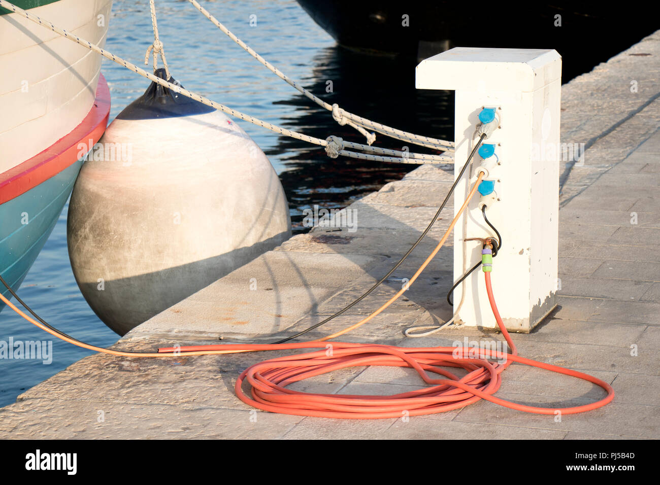 Pier with electrical and water supply pedestal with hose and cable , bitt and a rope on the dock fastened to a vintage boat with fender Stock Photo