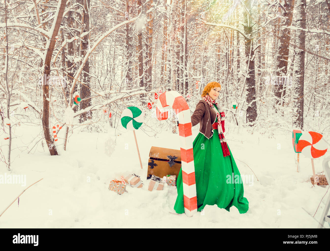 fiery red-haired woman in a ball green dress with a red leather belt in the costume of dwarf assistant Santa Claus in the winter forest with huge candy, a chest of gifts, the concept of the new year Stock Photo