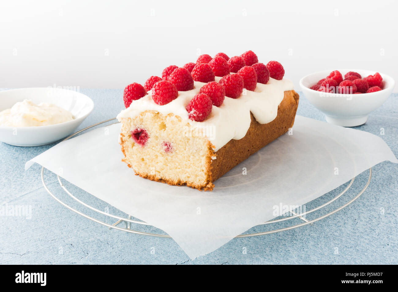 Closeup of a homemade raspberry loaf cake on parchment paper and cooling rack. Bowls filled with fresh raspberries and frosting in the background. Stock Photo