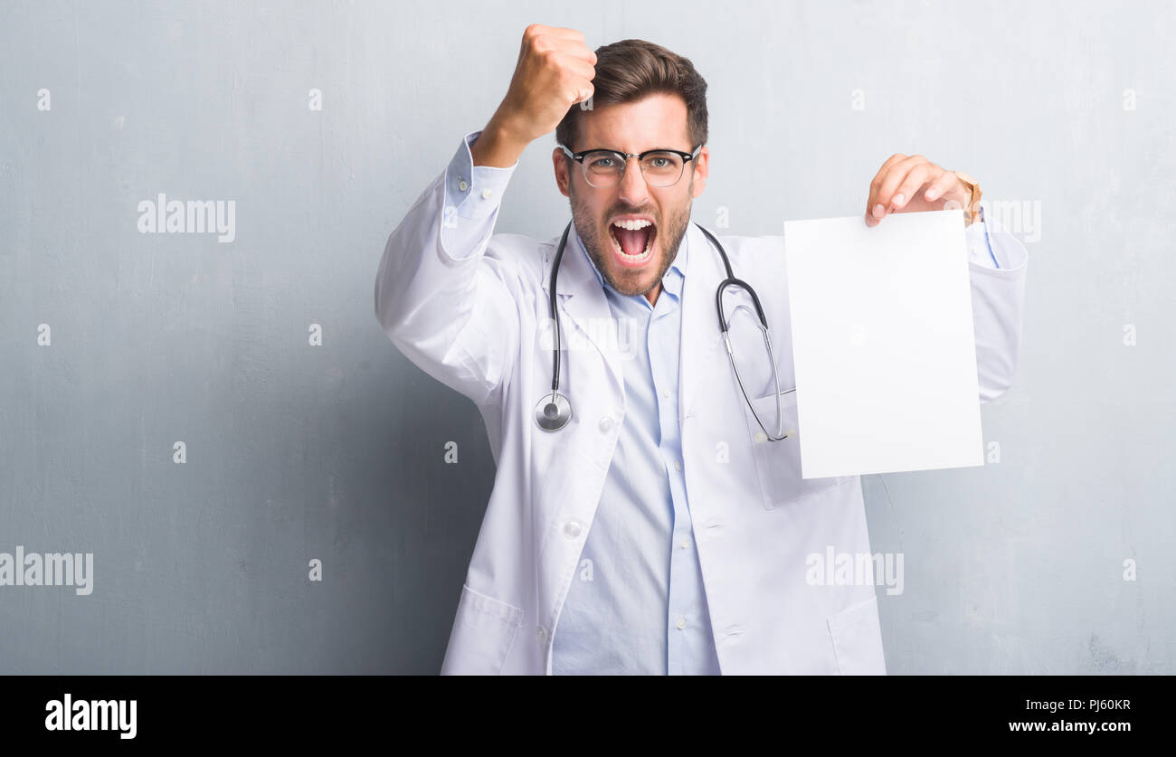 Handsome young doctor man over grey grunge wall holding blank sheet ...