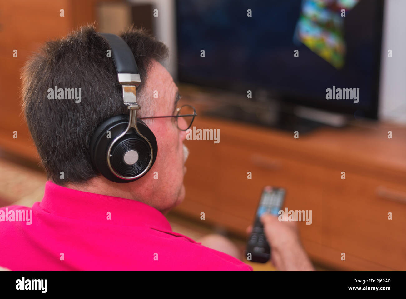 Back view of senior man with headphones watching tv holding remote control Stock Photo