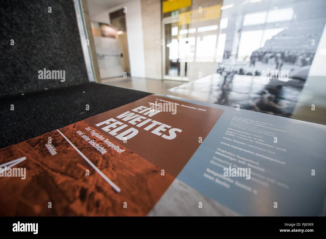04.09.2018, Berlin: A poster with the inscription 'Ein weites Feld. Tempelhof Airport and its History' (lit. A wide field - Tempelhof Airport and its history) is located next to a glass pane in the Tempelhof airport building. In the exhibition 'A wide field - Tempelhof Airport and its history' in the departure terminal, visitors can obtain information about the history of the airport on the basis of photos. The exhibition is open to visitors from 5 September to 30 December 2018. Photo: Wolfgang Kumm/dpa Stock Photo