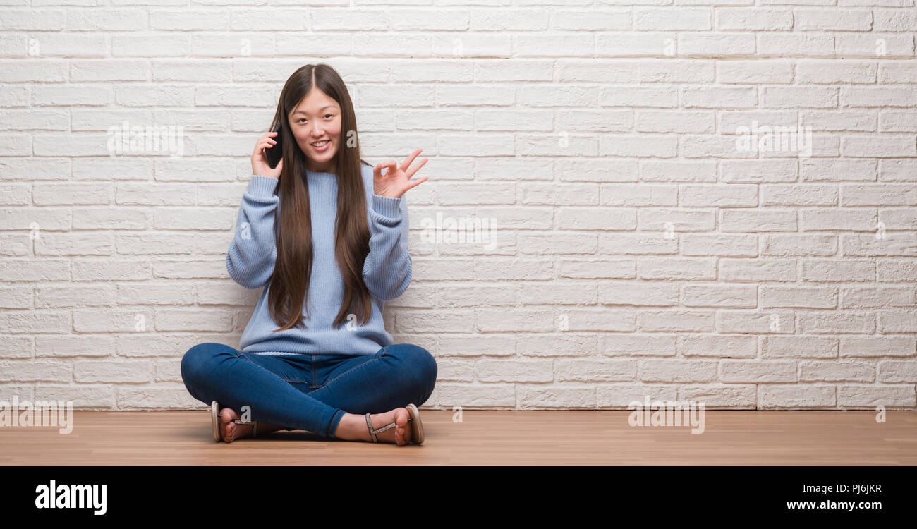 Young Chinese woman sitting on the floor over brick wall doing ok sign with fingers, excellent