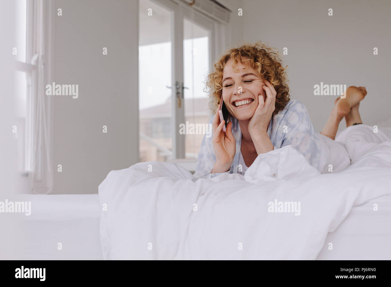 Smiling woman talking on mobile phone lying in bed after waking up in the morning. Stock Photo