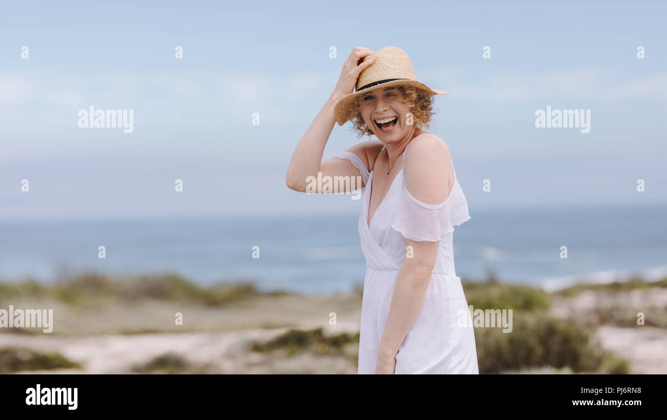 Smiling woman wearing a hat standing with sea in the background. Cheerful woman in white gown laughing holding her hat. Stock Photo