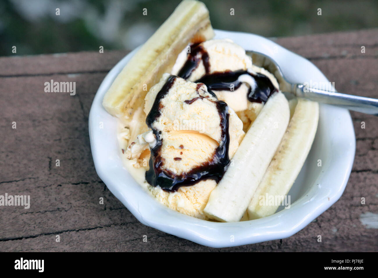 Hard ice cream just starting to melt, drizzled with chocolate sauce and garnished with banan slices in a white bowl on a painted brown wood surface wi Stock Photo