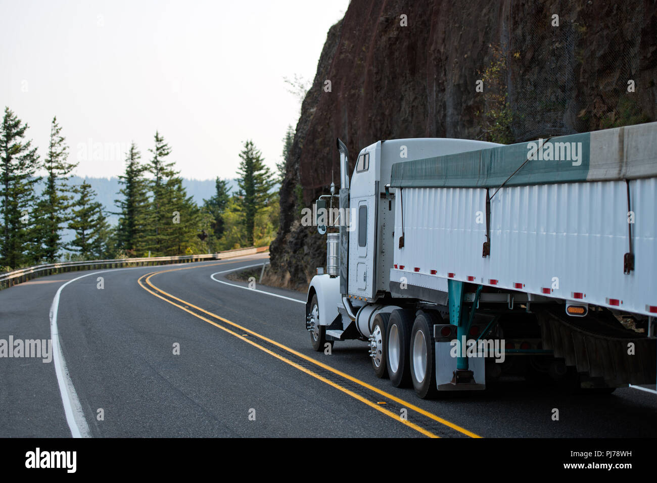 Big rig white American model bonnet semi truck tractor with plastic covered  long bulk semi trailer driving on the curve mountain road with rock mounta  Stock Photo - Alamy