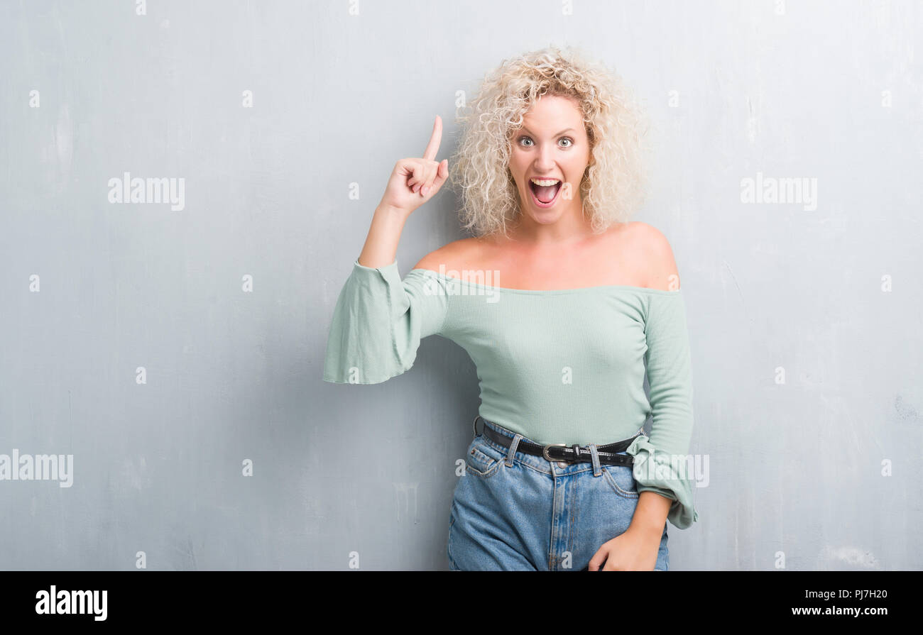 Young blonde woman with curly hair over grunge grey background pointing ...