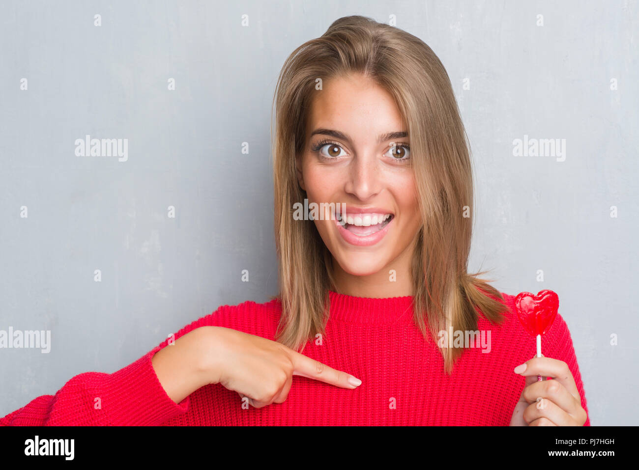 Beautiful young woman over grunge grey wall eating red heart lollipop
