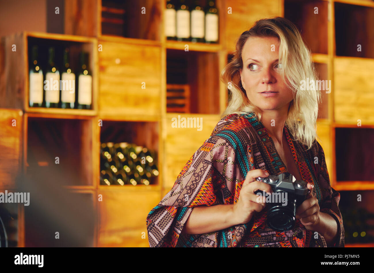 Portrait of a young pensive blond woman holding photo camera and looking away, wine chiller cabinet on a background. Photographer female posing and ph Stock Photo