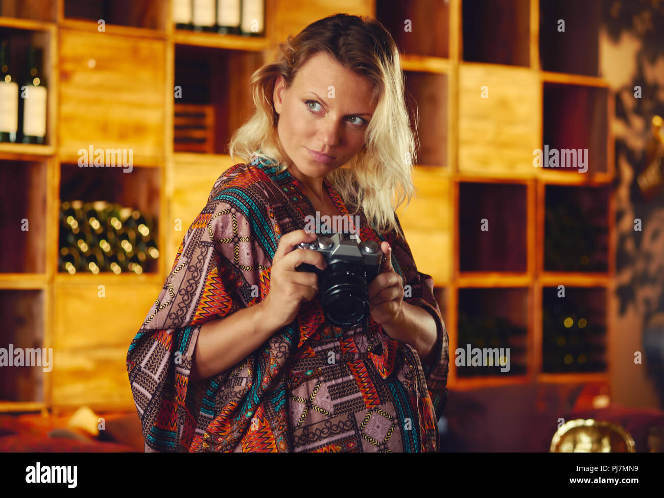 Portrait of a young pregnant pensive blond woman holding photo camera and looking away, wine chiller cabinet on a background. Photographer female posi Stock Photo