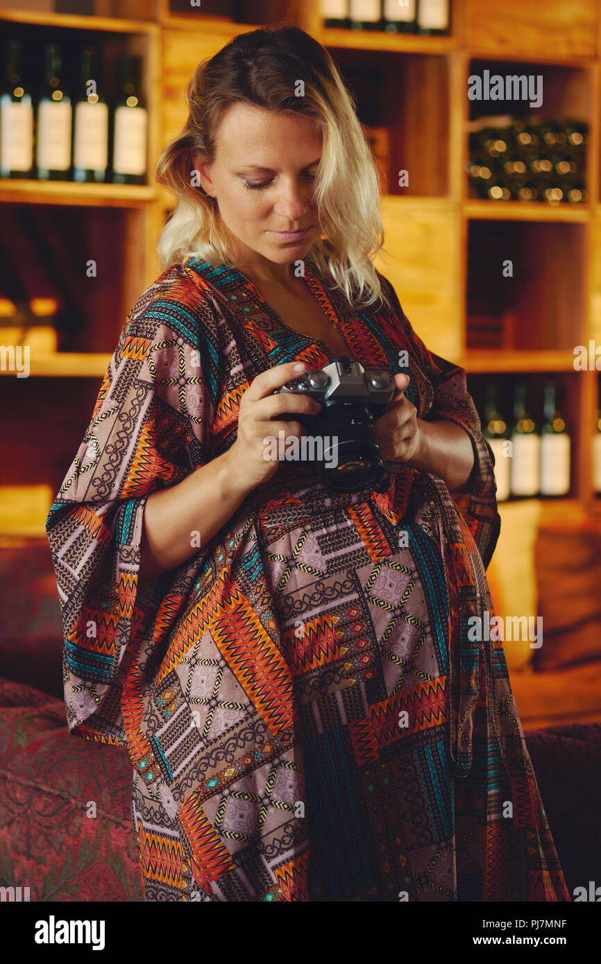 Portrait of a young pregnant pensive blond woman holding and looking on photo camera, wine chiller cabinet on a background. Photographer female posing Stock Photo