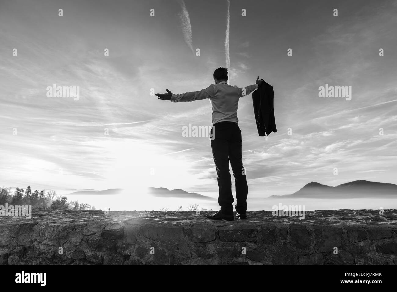 Monochrome image of successful business man at sunrise standing against the bright glow of the sun with outstretched arms, rear view. Stock Photo