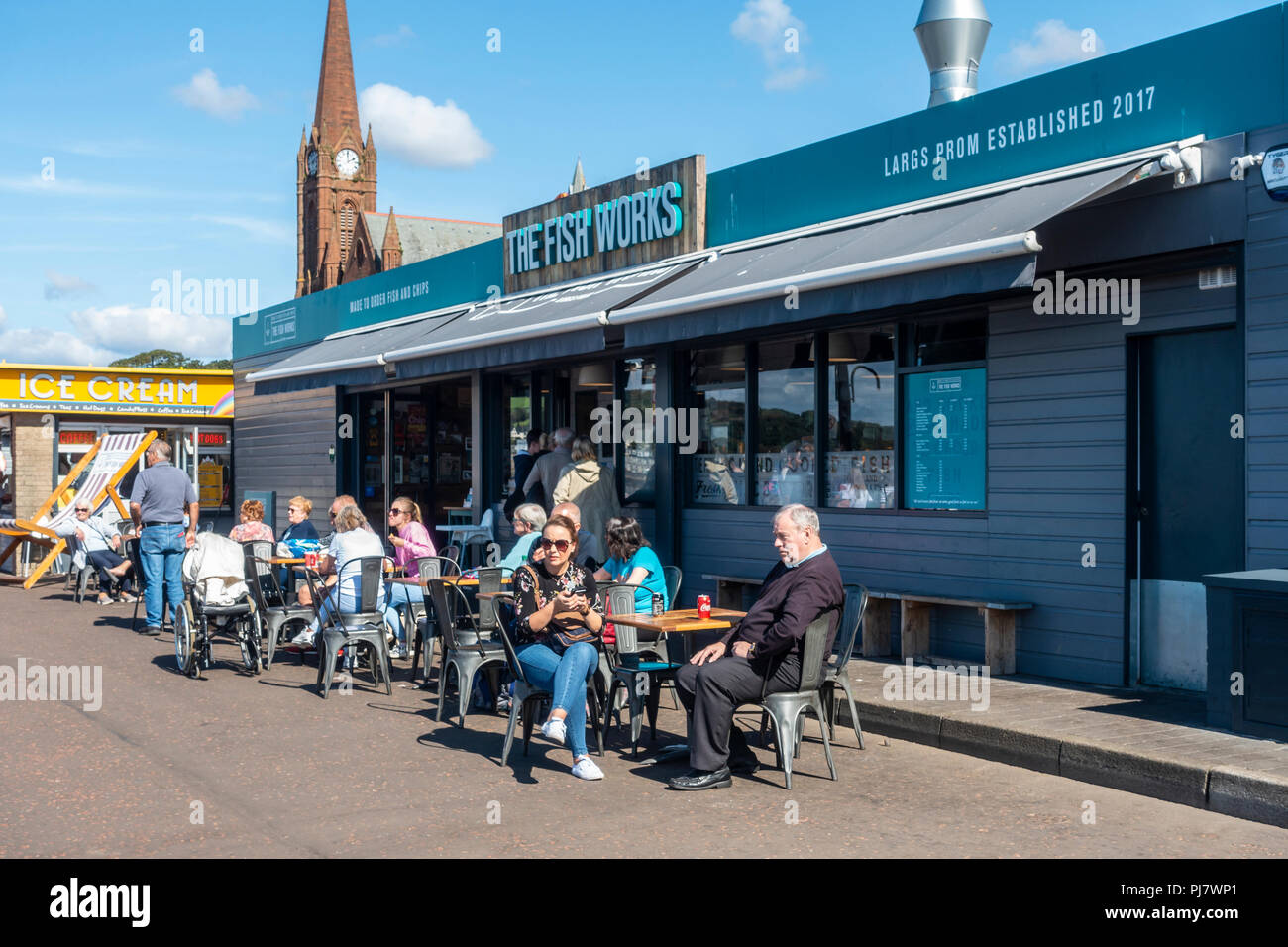 Customers eating and drinking outside The Fish Works, a fish and chips shop on Largs Promenade, North Ayrshire, on a sunny early September day. Stock Photo