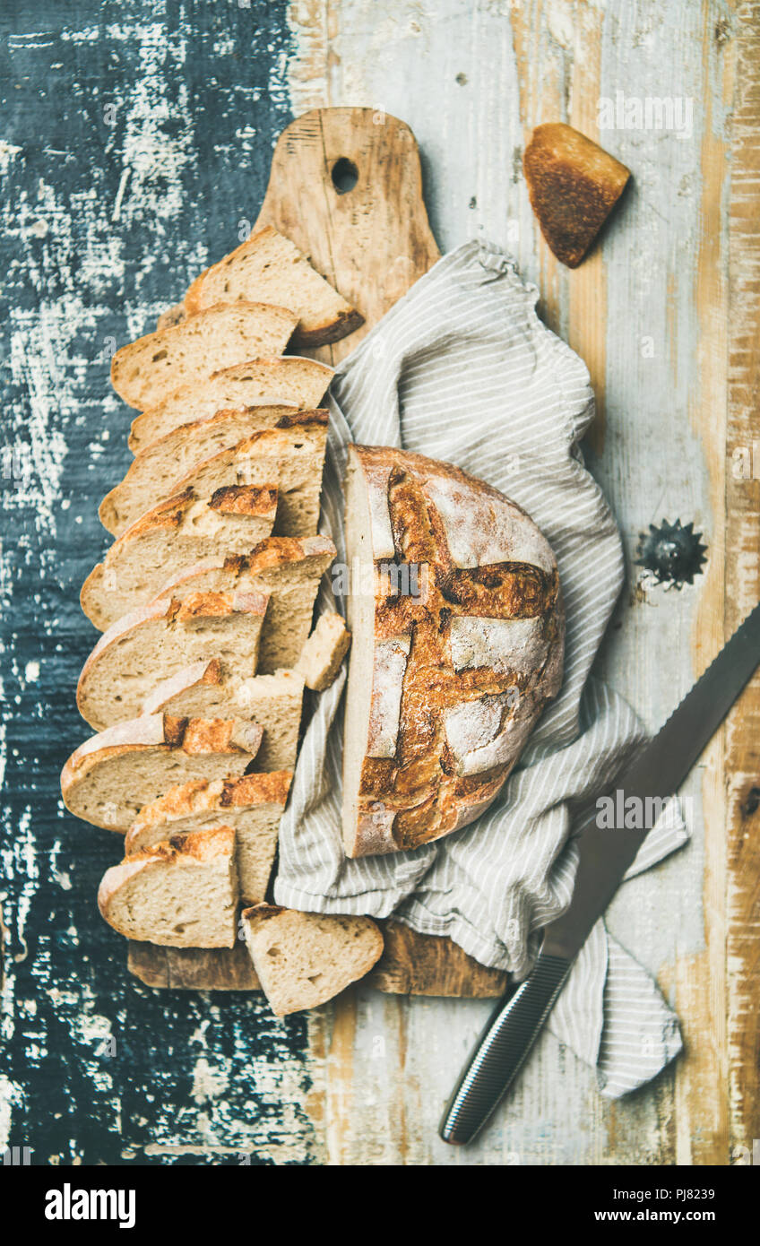 Flat-lay of freshly baked sourdough wheat bread loaf halved and cut in slices on board over linen napkin and rustic wooden table background, top view Stock Photo