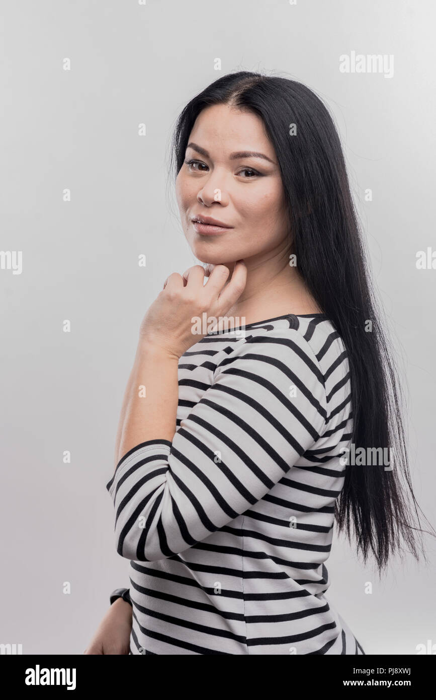 Dark-eyed woman with long straight hair standing in front of white wall ...