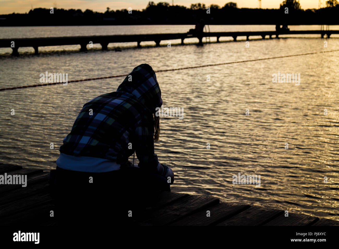Young sad female girl sitting on wooden pier looking at the water Stock Photo