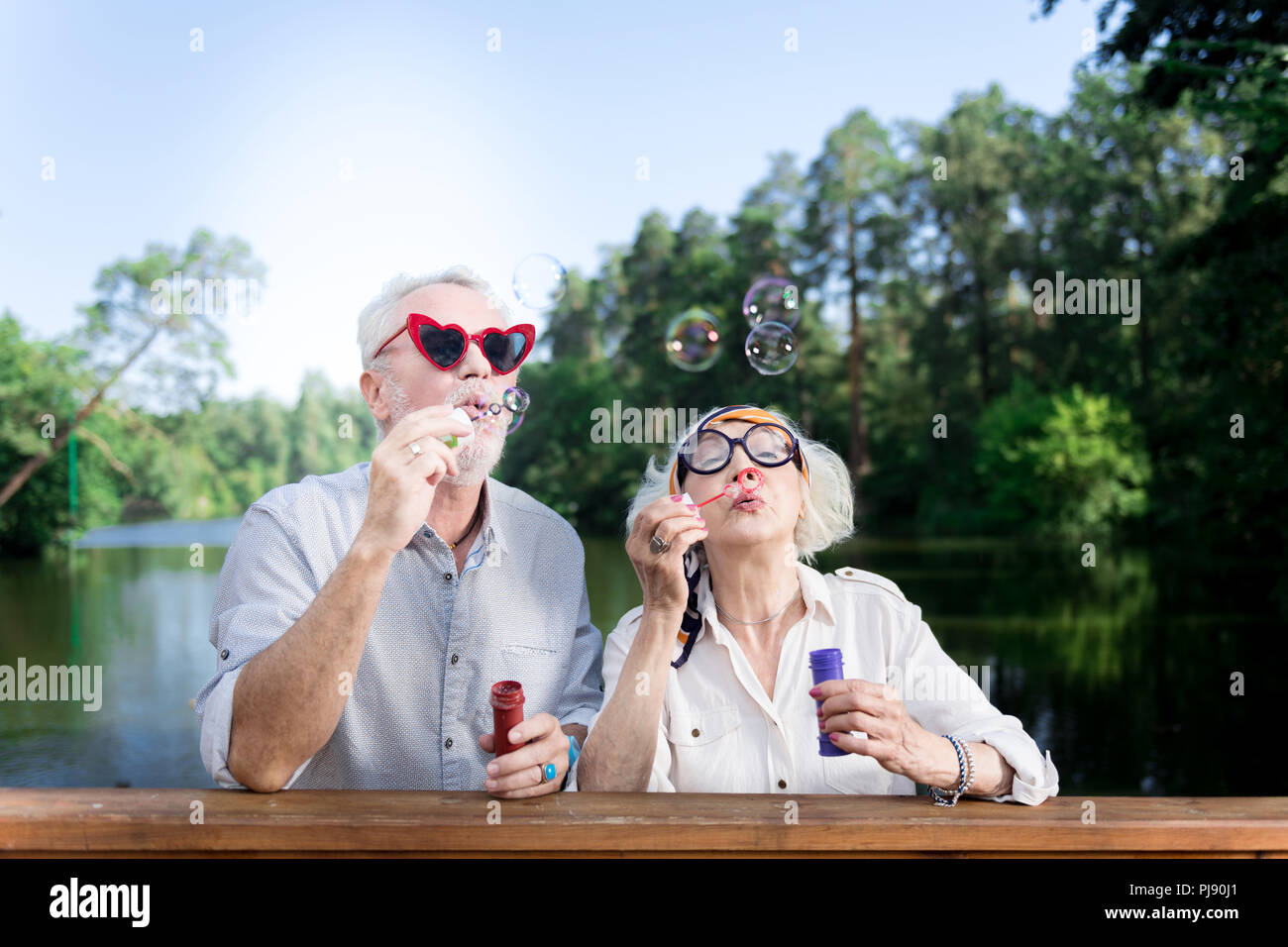 Funny elderly man wearing red heart shape sunglasses using soap bubbles Stock Photo
